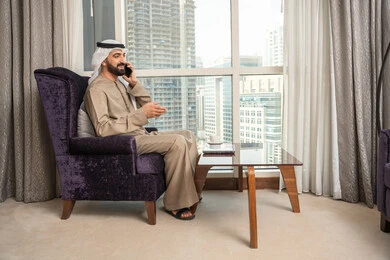 إيماءات السعادة بالتواصل مع الأصدقاء والأقارب، إجراء مكالمة هاتفية صوتية عبر الجوال، الاستمتاع بتبادل الأحاديث و الأخبار، صورة من الجانب لرجل عربي اماراتي خليجي يرتدي الكندورة والغترة الاماراتية يجلس في غرفة المعيشة بجانب النافذة يتحدث عبر الهاتف المحمول
