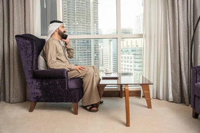 إجراء مكالمة هاتفية صوتية عبر الجوال، الاستمتاع بتبادل الأحاديث و الأخبار، صورة من الجانب لرجل عربي اماراتي خليجي يرتدي الكندورة والغترة الاماراتية يجلس في غرفة المعيشة بجانب النافذة يتحدث عبر الهاتف المحمول، إيماءات السعادة بالتواصل مع الأصدقاء والأقارب