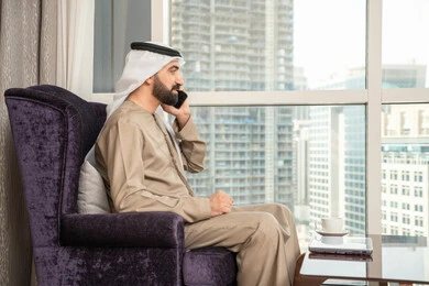 الاستمتاع بتبادل الأحاديث و الأخبار، إجراء مكالمة هاتفية صوتية عبر الجوال،صورة من الجانب لرجل عربي اماراتي خليجي يرتدي الكندورة والغترة الاماراتية يجلس في غرفة المعيشة بجانب النافذة يتحدث عبر الهاتف المحمول، إيماءات السعادة بالتواصل مع الأصدقاء والأقارب
