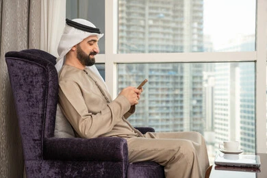 العمل عن بعد، تصفح التطبيقات وبرامج التواصل الاجتماعي، تبادل الأحاديث والتواصل والأخبار مع الأهل والأصدقاء، صورة من الجانب لرجل عربي خليجي اماراتي يرتدي الكندورة و الغتره يجلس بجانب النافذة يمسك بيده الهاتف المحمول، استخدام جهاز تقني حديث ومتطور
