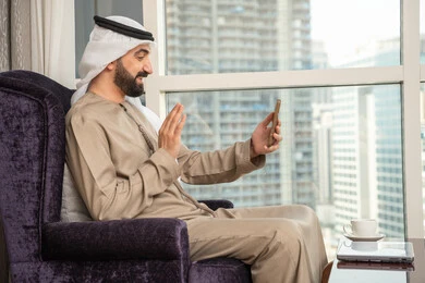 رفع كف اليد بتعابير السلام و التحية، الدردشة والتحدث مع الأصدقاء، استخدام التقنيات والتكنولوجيا الحديثة بالحياة اليومية ، اجراء مكالمه فيديو، تصفح مواقع وتطبيقات الانترنت،  صورة من الجانب لرجل عربي خليجي اماراتي يرتدي الكندوره والغتره يجلس بجانب النافذة يمسك الجوال بيده