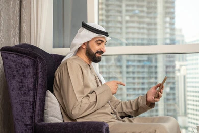 اجراء مكالمه فيديو ، استخدام التقنيات والتكنولوجيا الحديثة بالحياة اليومية ،الدردشة والتحدث مع الأصدقاء،تصفح مواقع وتطبيقات الانترنت،  صورة من الجانب لرجل عربي خليجي اماراتي يرتدي الكندوره والغتره يجلس بجانب النافذة يمسك الجوال بيده