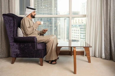Enjoying favorite drinks, sitting and enjoying the outdoor views through the window, a side image of an Emirati Gulf Arab man wearing a ghutrah and kandura sitting on a chair holding a cup of coffee in his hands, gestures of happiness and joy.