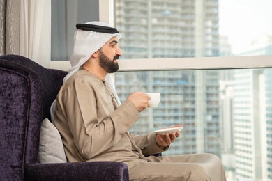 الجلوس والاستمتاع بالمناظر الخارجيه عبر النافذه، صورة من الجانب لرجل عربى خليجى اماراتى يرتدى الغترة و الكندورة يجلس على كرسي يمسك بيديه كوب من القهوة، الاستمتاع بتناول المشروبات المفضلة، ايماءات السعادة و السرور