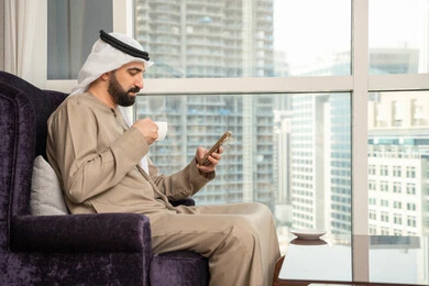تبادل الأحاديث والتواصل والأخبار مع الأهل والأصدقاء، صورة من الجانب لرجل عربي خليجي اماراتي يرتدي الكندورة و الغتره يجلس بجانب النافذة يمسك بيده الهاتف المحمول، استخدام جهاز تقني حديث ومتطور، العمل عن بعد، تصفح التطبيقات وبرامج التواصل الاجتماعي, تناول المشروب المفضل