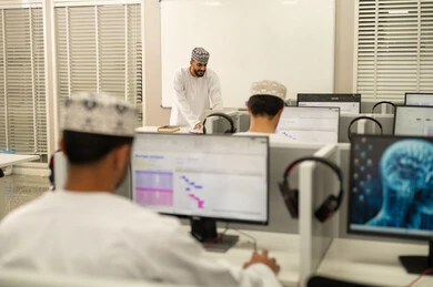 التعلم الرقمي للطلبة الجامعيين، مهنة المعلم في الجامعات التعليمية، اقامة دورات تدريبية في عمان، معلم عربي خليجي عماني يرتدي الدشداشة و الكمة يقف امام السبورة، صورة من الخلف لمجموعه من الطلاب العمانيون يجلسون امام الحاسوب يستمعون الى شرح الدرس