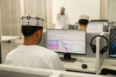 University study via computer, the profession of a teacher in educational universities, holding training courses in Oman, an Arab Gulf Omani teacher wearing a dishdasha and kumma standing in front of the blackboard, a picture from behind of a group of Omani students sitting in front of the computer listening to the lesson explanation.