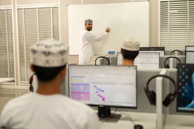 كتابة المعلومات على البورد الأبيض، مهنة المعلم في الجامعات التعليمية، اقامة دورات تدريبية في عمان، معلم عربي خليجي عماني يرتدي الدشداشة و الكمة يقوم بالكتابة على السبورة، مجموعه من الطلاب العمانيون يجلسون امام الحاسوب يستمعون الى شرح الدرس