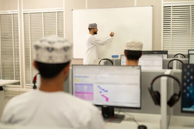 استخدام البورد لتدوين الملاحظات، مهنة المعلم في الجامعات التعليمية، اقامة دورات تدريبية في عمان، معلم عربي خليجي عماني يرتدي الدشداشة و الكمة يقوم بالكتابة على السبورة، مجموعه من الطلاب العمانيون يجلسون امام الحاسوب يستمعون الى شرح الدرس