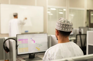 تسجيل المعلومات على البورد، مهنة المعلم في الجامعات التعليمية، اقامة دورات تدريبية في عمان، معلم عربي خليجي عماني يرتدي الدشداشة و الكمة يقوم بالكتابة على السبورة، مجموعه من الطلاب العمانيون يجلسون امام الحاسوب يستمعون الى شرح الدرس