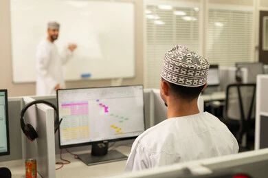 مهنة المعلم في الجامعات التعليمية، كتابة وتدوين الملاحظات على بورد ابيض، اقامة دورات تدريبية في عمان، معلم عربي خليجي عماني يرتدي الدشداشة و الكمة يقوم بالكتابة على السبورة، مجموعه من الطلاب العمانيون يجلسون امام الحاسوب يستمعون الى شرح الدرس