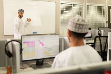 كتابة وتدوين الملاحظات على بورد ابيض، اقامة دورات تدريبية في عمان، معلم عربي خليجي عماني يرتدي الدشداشة و الكمة يقوم بالكتابة على السبورة، مهنة المعلم في الجامعات التعليمية، مجموعه من الطلاب العمانيون يجلسون امام الحاسوب يستمعون الى شرح الدرس