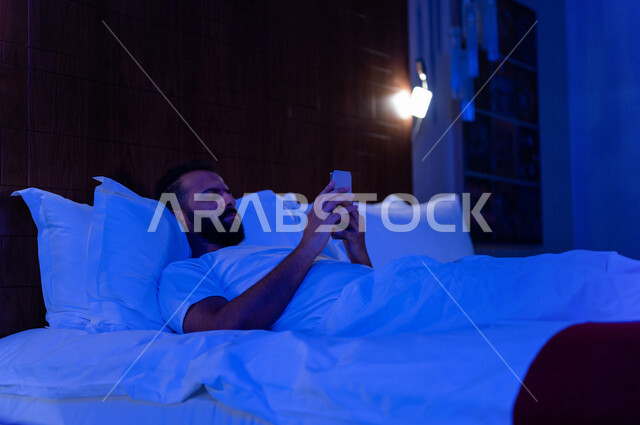 رجل عربي خليجي سعودي مستلقي على فراشه، يتصفح الانترنت من خلال الهاتف المحمول، الاسترخاء والراحة، استخدام تقنيات حديثة
