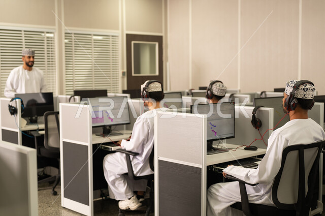 Using headphones while studying, academic universities in the Sultanate of Oman, a picture from behind of Omani students sitting in front of the computer listening to the lecture, an Arab Gulf university professor from Oman wearing a dishdasha and kumma explaining to his students in the lecture, advanced colleges and universities.