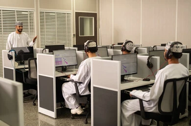 جامعات أكاديميه في سلطنه عمان ، استخدام تقنية الهيدفون، صورة من الجانب لطلاب عمانوين يجلسون امام الحاسوب يقومون بالإستماع الى المحاضره، أستاذ جامعي عربي خليجي عماني يرتدي الدشداشة و الكمة يشرح لطلابه في المحاضره, كليات وجامعات متطوره