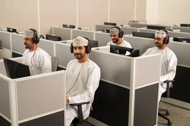 التعلم التفاعلي بالحاسوب الجامعي، مجموعة من الطلاب العربيون الخليجيون العمانوين يرتدون الدشداشة والكمه و سماعات الرأس يجلسون أمام أجهزة الكمبيوتر في مختبر حديث، التركيز على الدراسة والتفاعل الأكاديمي