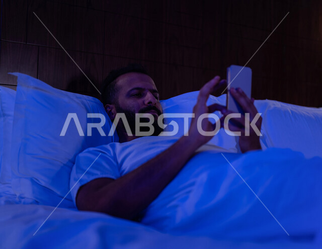 رجل عربي خليجي سعودي مستلقي على فراشه، يتصفح الانترنت من خلال الهاتف المحمول، الاسترخاء والراحة، استخدام تقنيات حديثة
