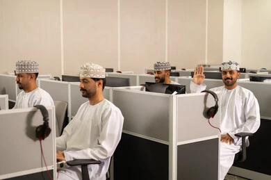 Learning through university devices, a group of Arab Gulf Omani students wearing dishdasha and kumma are sitting in front of computers in a modern lab, focusing on studying and academic interaction.