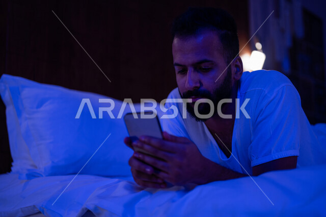 رجل عربي خليجي سعودي مستلقي على فراشه، تصفح الانترنت من خلال الهاتف المحمول، اللعب والترفيه، استخدام تقنيات حديثة