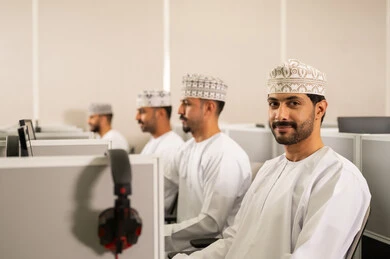 التعلم الرقمي في الجامعة، مجموعة من الطلاب العربيون الخليجيون العمانوين يرتدون الدشداشة والكمه يجلسون أمام أجهزة الكمبيوتر في مختبر حديث، التركيز على الدراسة والتفاعل الأكاديمي