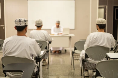 التعليم الجامعي  في سلطنه عمان، الانخراط والمشاركة خلال المحاضرة، مجموعه من الطلاب يستمعون الى الدرس بايماءات الاندماج و التركيز، معلم عربي خليجي عماني يرتدي الدشداشة و الكمة يقوم بشرح الدرس للطلاب