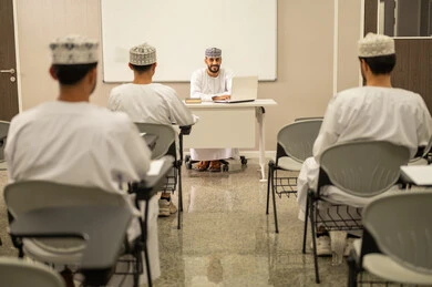 Positive interaction during the lesson, a group of students listening to the lesson with gestures of engagement and focus, an Omani Gulf Arab teacher wearing the dishdasha and kumma explaining the lesson to the students, education in the Sultanate of Oman.