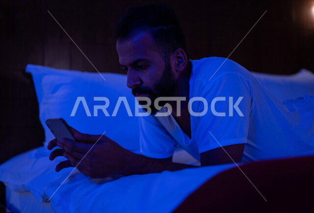 رجل عربي خليجي سعودي مستلقي على فراشه، تصفح الانترنت من خلال الهاتف المحمول، اللعب والترفيه، استخدام تقنيات حديثة