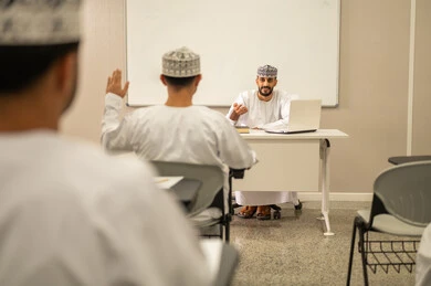 رفع كف اليد للاجابة على السؤال، مجموعه من الطلاب يستمعون الى الدرس بايماءات الاندماج و التركيز، التفاعل مع المعلم و التلاميذ أثناء المحاضرات الدراسية، معلم عربي خليجي عماني يرتدي الدشداشة و الكمة يقوم بشرح الدرس للطلاب، التعليم في سلطنه عمان