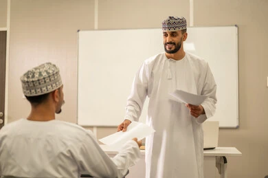 الاجتهاد في الدراسة للوصول إلى النجاح, مجموعة من التلاميذ العمانيين يقومون بالاستعداد لحل أسئلة الامتحان, اهداف التربية و التعليم, التعليم في سلطنه عمان , معلم عربي خليجي عماني يرتدي الدشداشة و الكمه يقوم بتوزيع ورق الامتحان على الطلاب