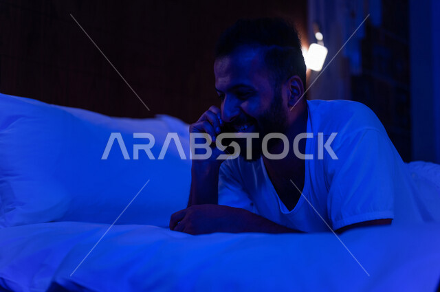 رجل عربي خليجي سعودي مستلقي على فراشه، يجري اتصال من خلال الهاتف المحمول، التواصل الاجتماعي والدردشة، استخدام تقنيات حديثة، ايماءات وجه سعيدة