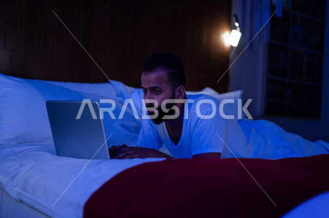 رجل عربي خليجي سعودي مستلقي على فراشه، انجاز الاعمال من خلال استخدم جهاز الحاسوب المحمول، العمل عن بعد، استخدام تقنيات حديثة