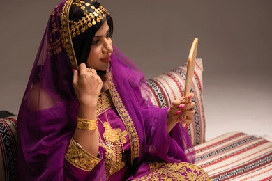 مفهوم التزيين والإطلالة المتألقة، الاحتفالات والمناسبات السعيدة ،التزين بالحلي و المجوهرات الذهبية ، استخدام مساحيق التجميل لإبراز الجمال، عروس عربية خليجية اماراتية ترتدي الزي التقليدي الشعبي تنظر لنفسها بالمرآة، الاستعداد للاحتفال بليلة العمر