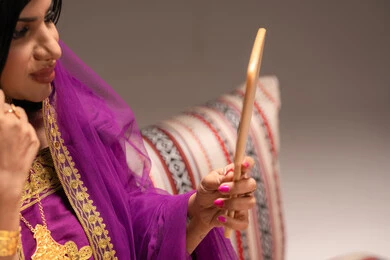 الاحتفالات والمناسبات السعيدة ، مفهوم التزيين والإطلالة المتألقة،التزين بالحلي و المجوهرات الذهبية ، استخدام مساحيق التجميل لإبراز الجمال، صورة مقربة لعروس عربية خليجية اماراتية ترتدي الزي التقليدي الشعبي تنظر لنفسها بالمرآة، الاستعداد للاحتفال بليلة العمر