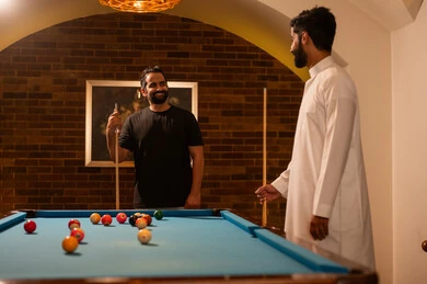 Discussion and dialogue among friends, billiard café, youth activities and hobbies, billiard table (snooker), a group of young Arab Saudi Gulf men wearing casual and traditional attire, playing billiards, professional players, competitions and challenges, fun and entertainment, youth sports hall.