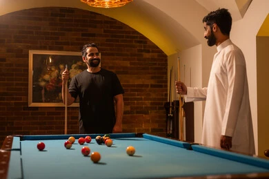 Professional players, dialogue and discussion among friends, billiard café, youth activities and hobbies, billiard table (snooker), a group of young Arab Saudi Gulf men wearing casual and traditional attire, playing billiards, competitions and challenges, fun and entertainment, youth sports hall.