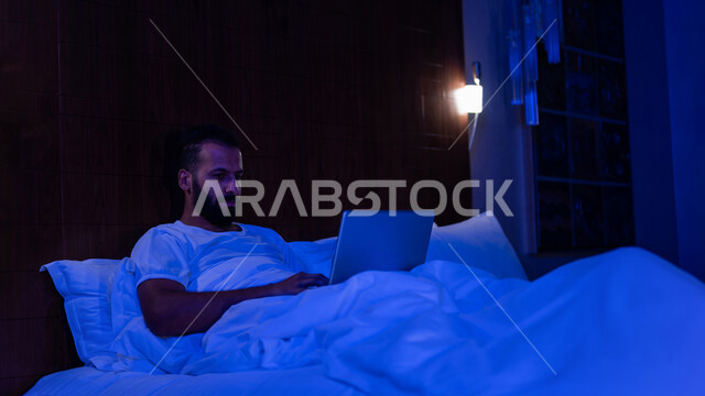 A Saudi Arabian Gulf man lying on his bed, doing business by using his laptop, working remotely, surfing the Internet, using modern technologies