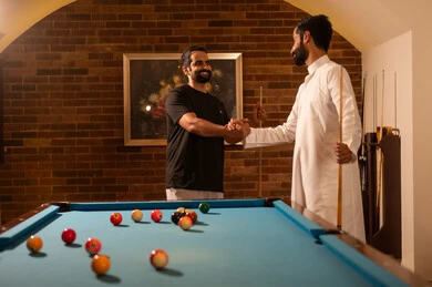 Playing and sportsmanship among friends, professional players, dialogue and discussion among friends, billiard café, youth activities and hobbies, billiard table (snooker), a group of young Arab Saudi Gulf men wearing casual and traditional attire, playing billiards, youth sports hall.