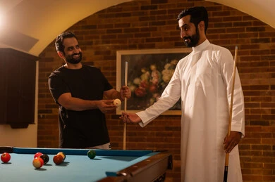 Cooperation and competition together, professional players, dialogue and discussion among friends, billiard café, youth activities and hobbies, billiard table (snooker), a group of young Arab Saudi Gulf men wearing casual and traditional attire, playing billiards, youth sports hall.