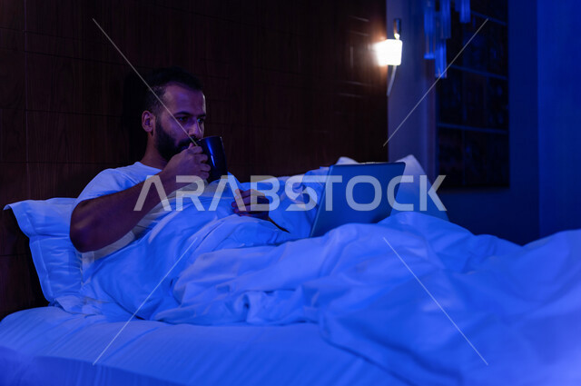 رجل عربي خليجي سعودي مستلقي على فراشه، انجاز الاعمال من خلال استخدم جهاز الحاسوب المحمول، العمل عن بعد، تصفح الانترنت، استخدام تقنيات حديثة