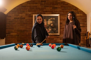 The passion for playing snooker, professional players, dialogue and discussion among friends, billiard café, youth activities and hobbies, billiard table (snooker), a group of Arab Saudi Gulf women wearing abayas, one of them wearing a hijab, playing billiards, youth sports hall.