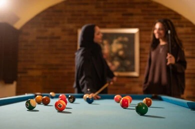 Two friends are talking in front of the game table, professional players, a dialogue and discussion among friends, a billiards café, youth activities and hobbies, a billiards table (snooker), a group of Arab Saudi Gulf women wearing abayas, one of them wearing a hijab, playing billiards, a youth sports hall.