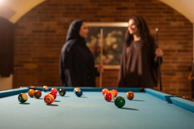 Expressions of joy and happiness, professional players, dialogue and discussion among friends, billiard café, youth activities and hobbies, billiard table (snooker), a group of Arab Saudi Gulf women wearing abayas, one of them wearing a hijab, playing billiards, youth sports hall.