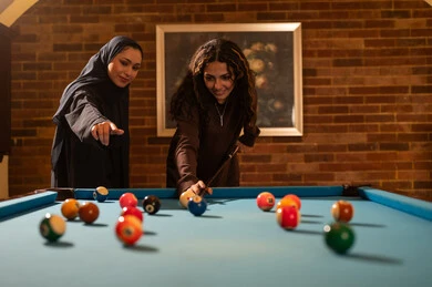 Elegant and modern rooms with a billiard table, professional players, conversation and discussion among friends, billiard café, youth activities and hobbies, billiard table (snooker), a group of Arab Saudi Gulf women wearing abayas, one of them wearing a hijab, playing billiards, youth gym.