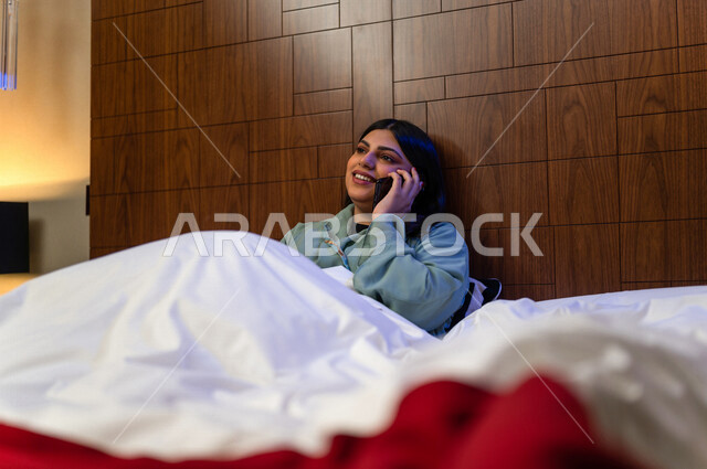 A Saudi Arabian Gulf woman lying on the bed, making a call through a mobile phone, social networking and chatting, using modern technologies, happy facial gestures