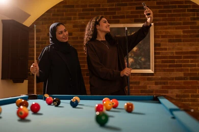 Taking a selfie, professional players, conversation and discussion among friends, billiard café, youth activities and hobbies, billiard table (snooker), a group of Arab Saudi Gulf women wearing abayas, one of them wearing a hijab, playing billiards, youth sports hall.