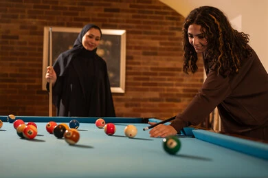 Playing, challenge, and competition, professional players, dialogue and discussion among friends, billiard café, youth activities and hobbies, billiard table (snooker), a group of Arab Saudi Gulf women wearing abayas, one of them wearing a hijab, playing billiards, youth sports hall.