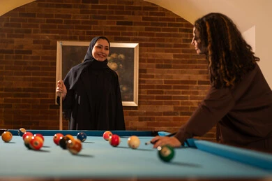 Focus, calmness, and aiming, professional players, dialogue and discussion among friends, billiard café, youth activities and hobbies, billiard table (snooker), a group of Arab Saudi Gulf women wearing abayas, one of them wearing a hijab, playing billiards, youth sports hall.
