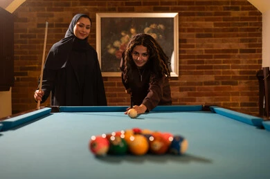 Competition and challenges among friends, professional players, dialogue and discussion among friends, billiard café, youth activities and hobbies, billiard table (snooker), a group of Arab Saudi Gulf women wearing abayas, one of them wearing a hijab, playing billiards, youth sports hall.