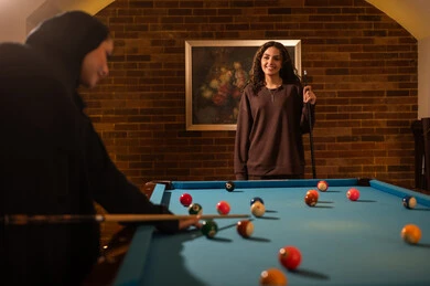 Smart play among friends, professional players, dialogue and discussion among friends, billiard café, youth activities and hobbies, billiard table (snooker), a group of Arab Saudi Gulf women wearing abayas, one of them wearing a hijab, playing billiards, youth sports hall.