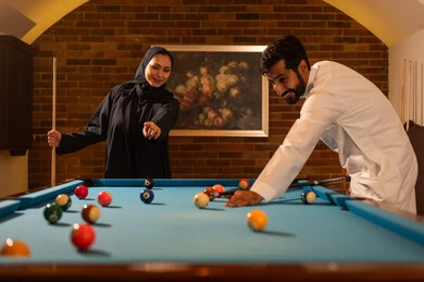 The concept of successful marital relationships, preferred times between couples, activities and youth hobbies, dialogue and discussion among friends, billiard café, billiard table (snooker), a man and his wife, Arab Saudi Gulf nationals wearing traditional attire, playing billiards, youth sports hall.